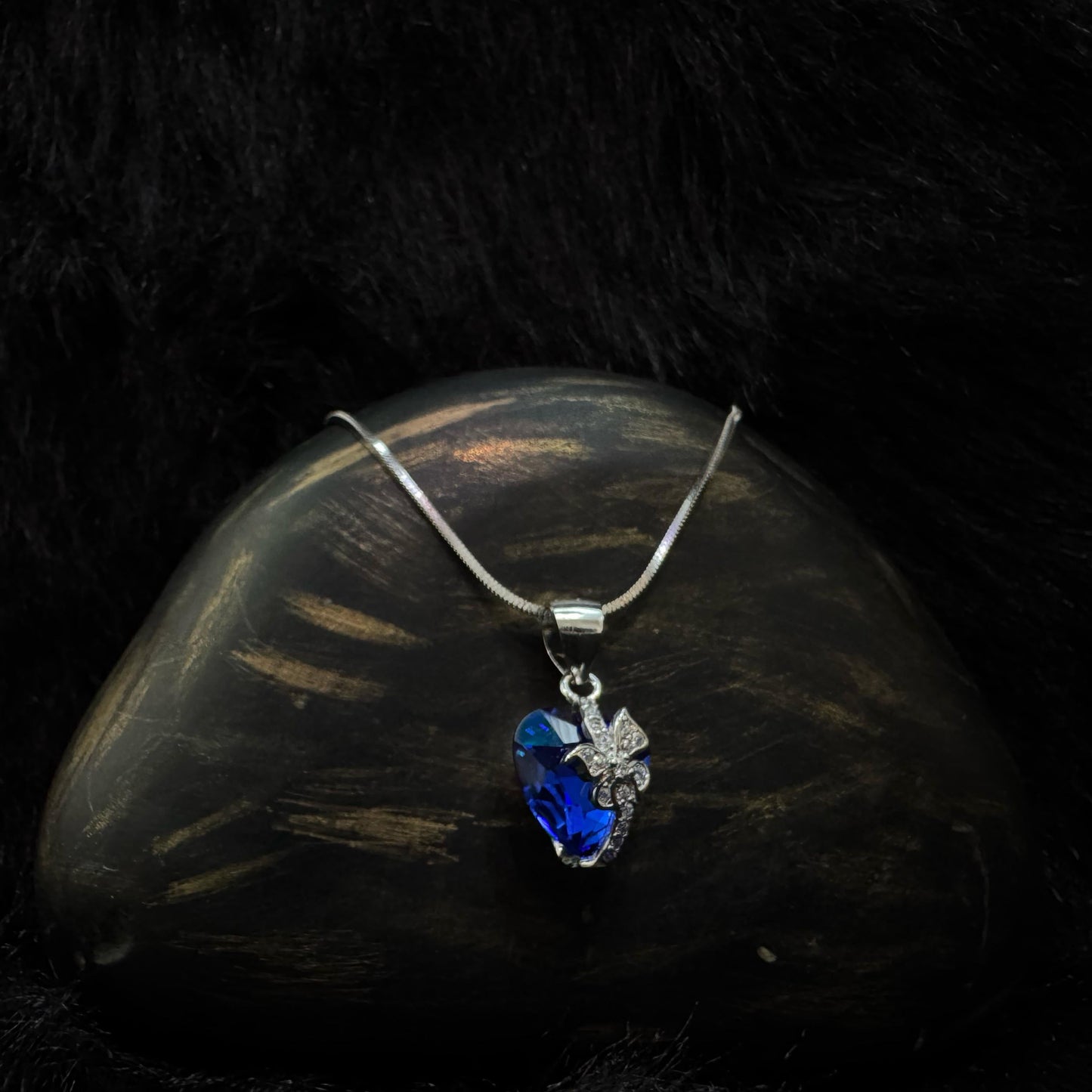 A silver necklace featuring a deep blue heart-shaped crystal pendant with a sparkling butterfly motif, displayed on a dark textured background.