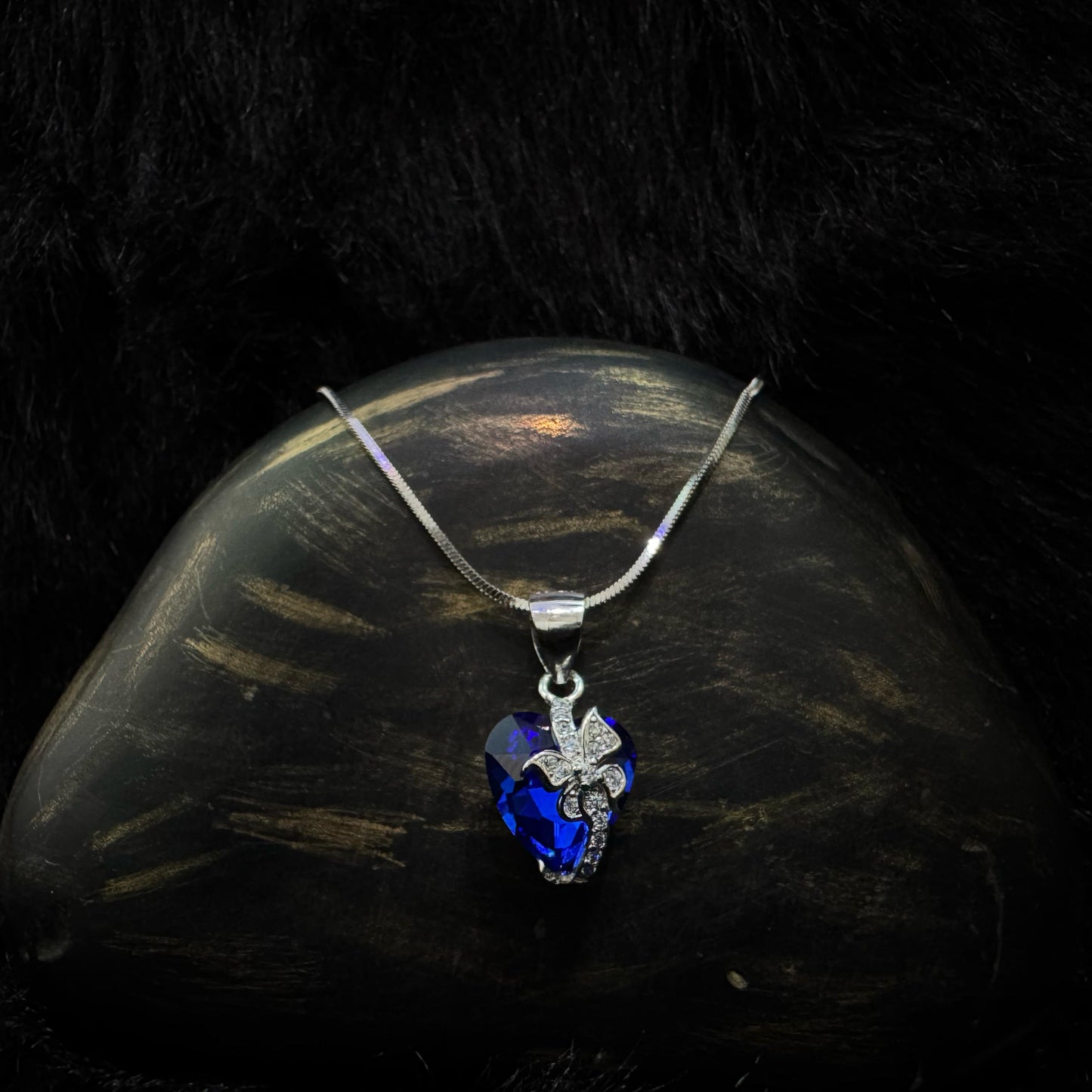 A silver necklace featuring a deep blue heart-shaped crystal pendant with a sparkling butterfly motif, displayed on a dark textured background.
