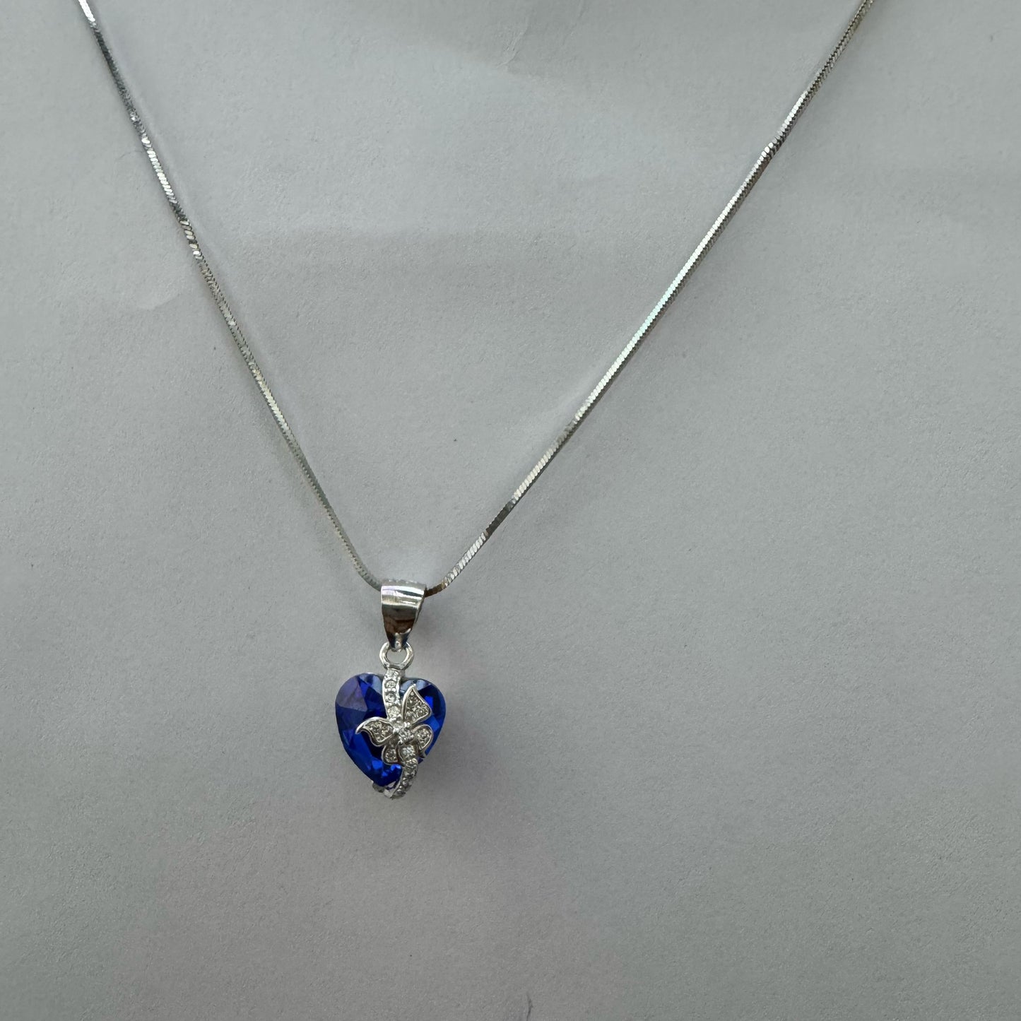 A silver necklace featuring a deep blue heart-shaped crystal pendant with a sparkling butterfly motif, displayed on a dark textured background.