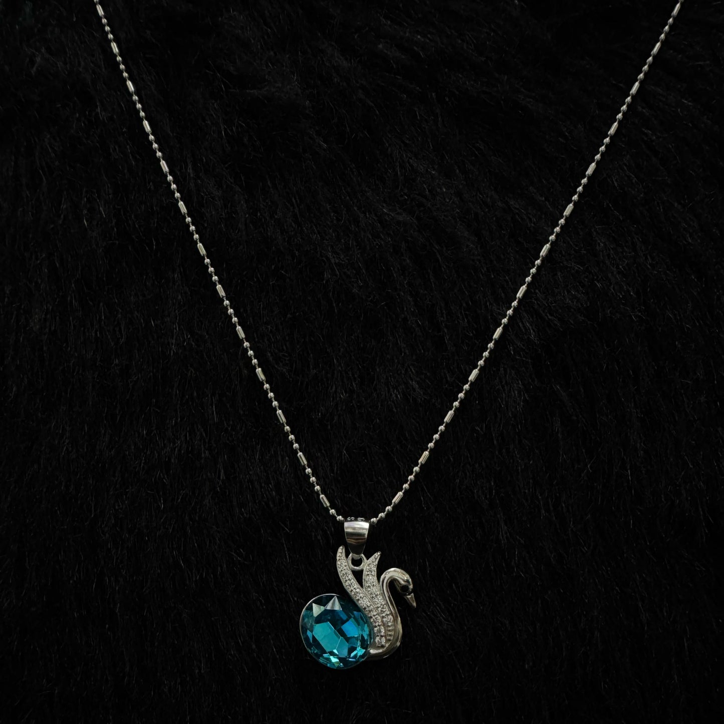 A silver necklace featuring an elegant swan motif adorned with cubic zirconia, holding a vivid blue oval-cut crystal, displayed against a textured black and gold background.