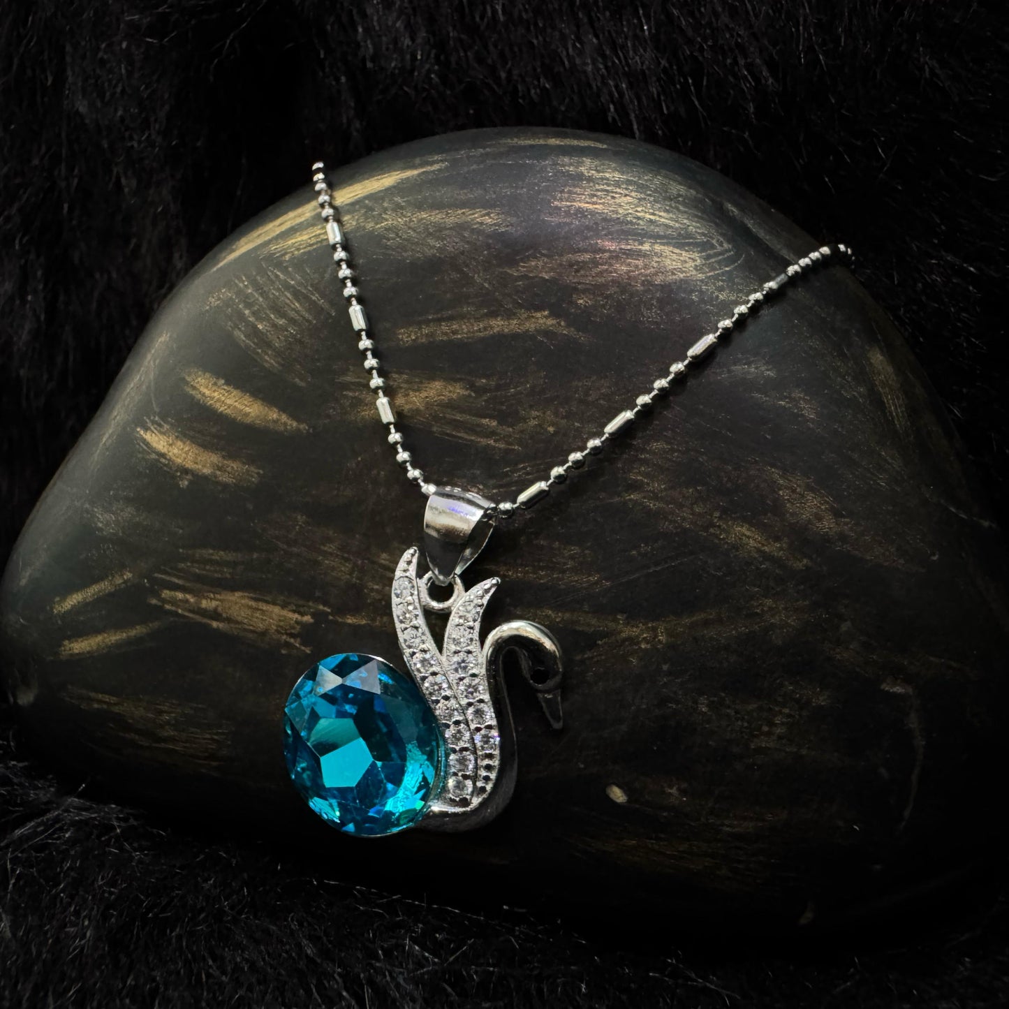 A silver necklace featuring an elegant swan motif adorned with cubic zirconia, holding a vivid blue oval-cut crystal, displayed against a textured black and gold background.