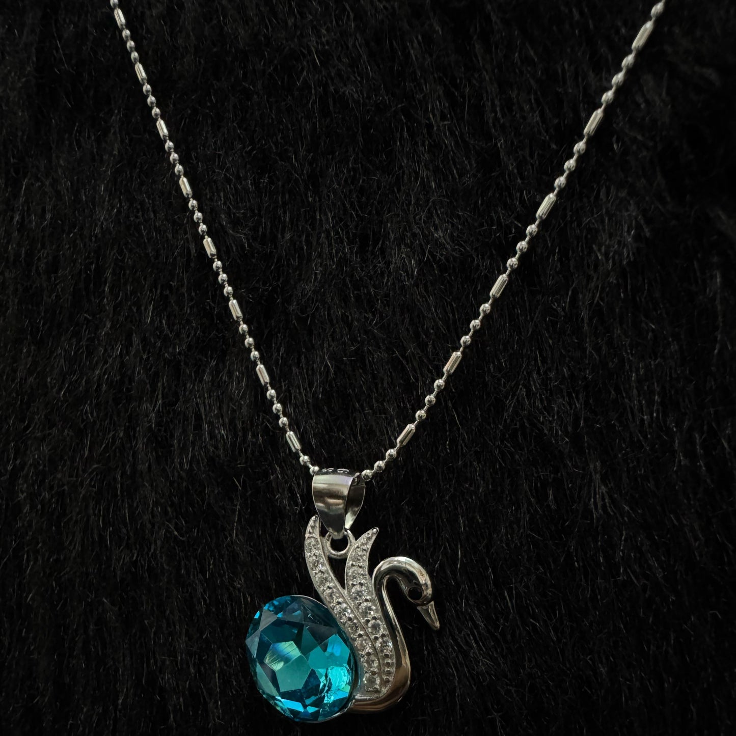 A silver necklace featuring an elegant swan motif adorned with cubic zirconia, holding a vivid blue oval-cut crystal, displayed against a textured black and gold background.