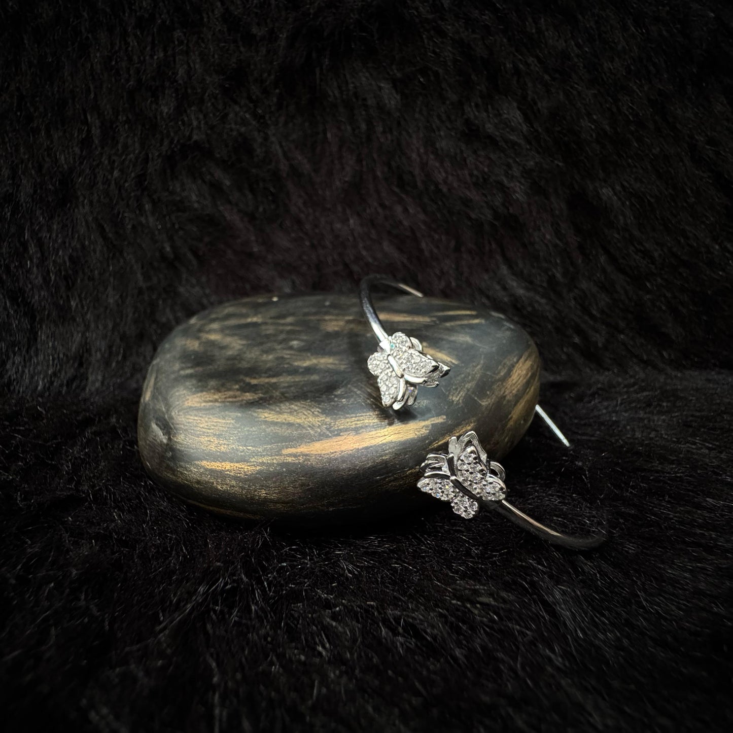 Elegant silver bangle bracelet with two butterfly-shaped ends adorned with sparkling cubic zirconia, displayed on a dark stone with a soft black background.