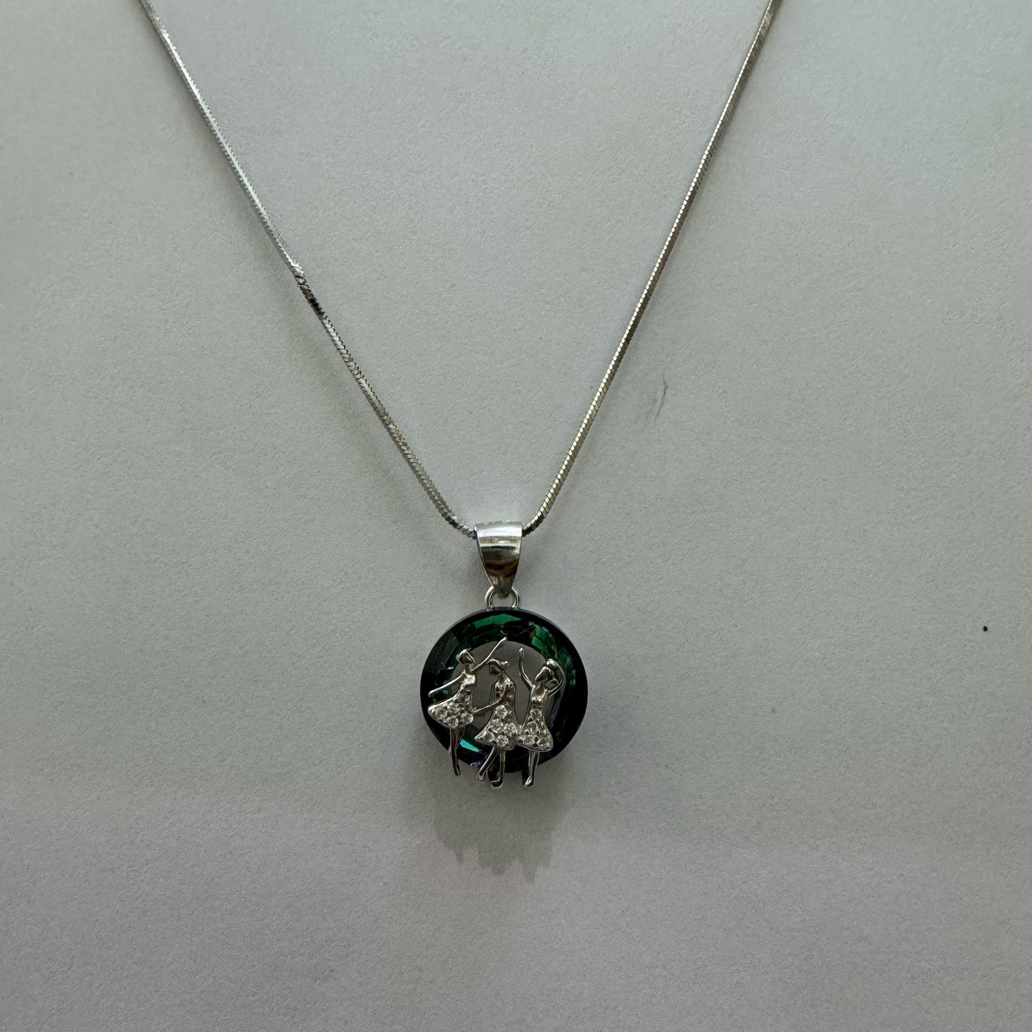 A sterling silver necklace with a circular pendant featuring two dancers adorned with cubic zirconia, set against a multicolor Swarovski crystal background, displayed on a textured black and gold surface.