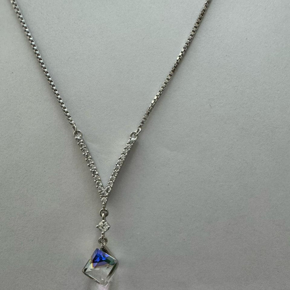 A silver rhinestone necklace featuring a bold cube-cut Swarovski crystal pendant, draped elegantly over a dark stone with a black background.