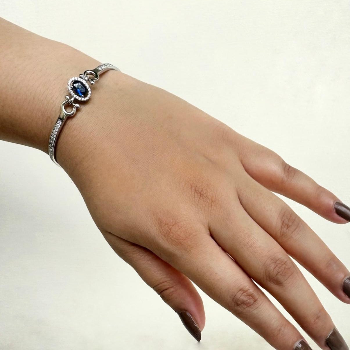 A hand wearing a 925 sterling silver bracelet featuring an oval sapphire-blue stone surrounded by small diamonds, photographed on a speckled white stone background.
