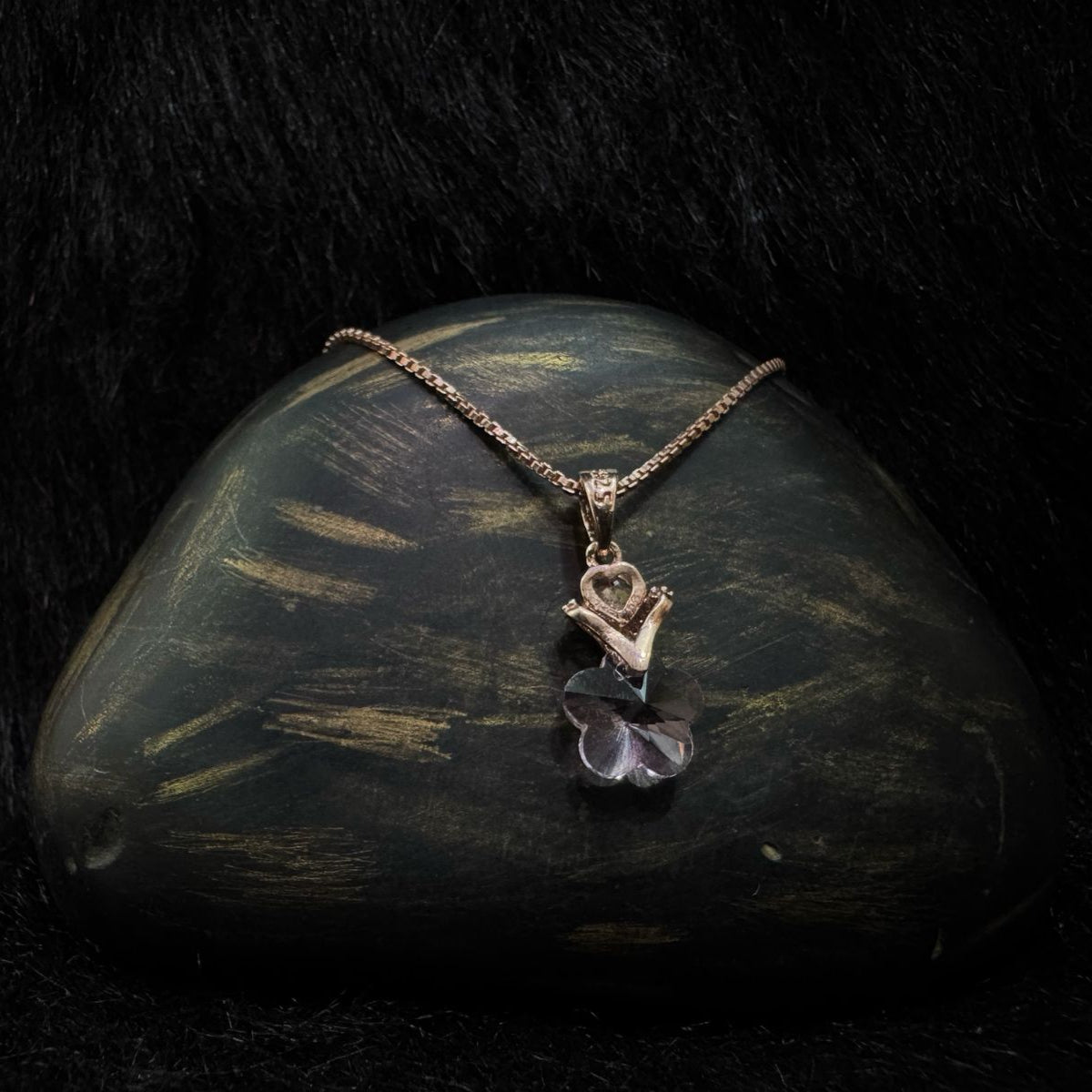 A rose gold necklace featuring a pendant of hands holding a heart above a clear faceted flower crystal, displayed elegantly on a dark stone with a black background.