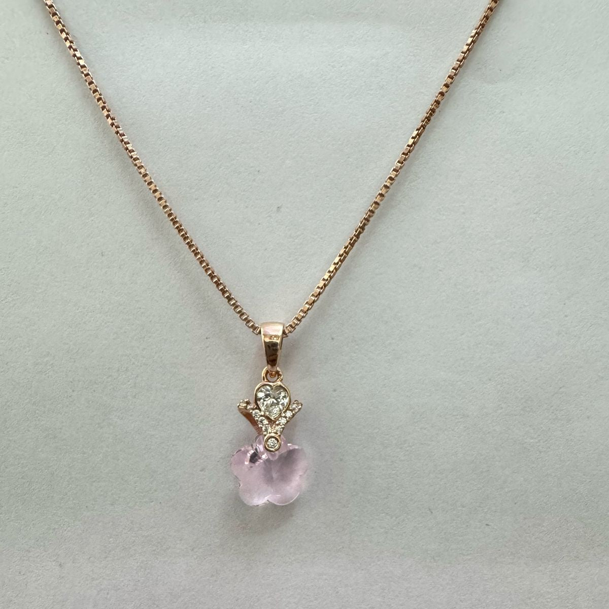 A rose gold necklace featuring a pendant of hands holding a heart above a clear faceted flower crystal, displayed elegantly on a dark stone with a black background.