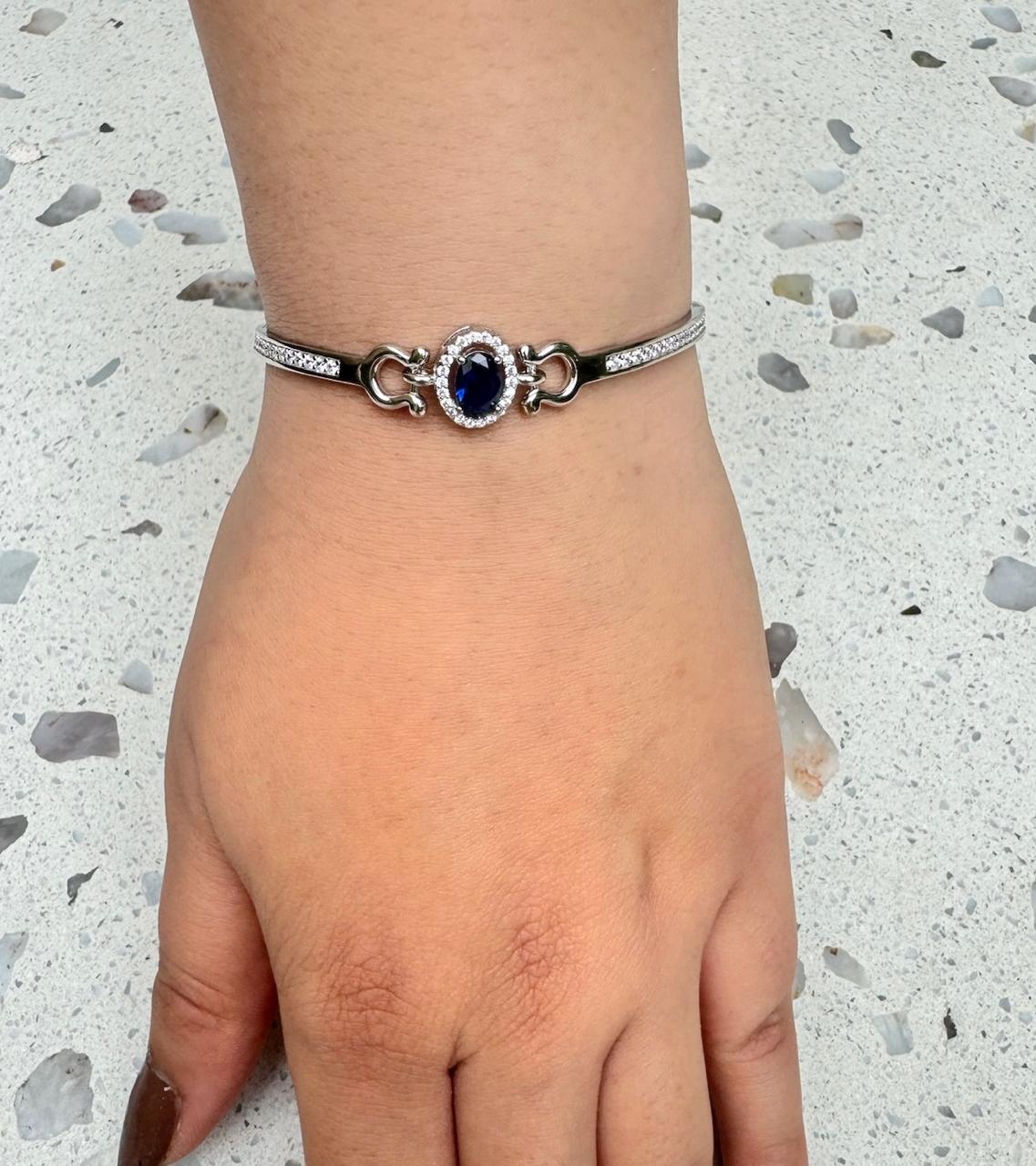A hand wearing a 925 sterling silver bracelet featuring an oval sapphire-blue stone surrounded by small diamonds, photographed on a speckled white stone background.