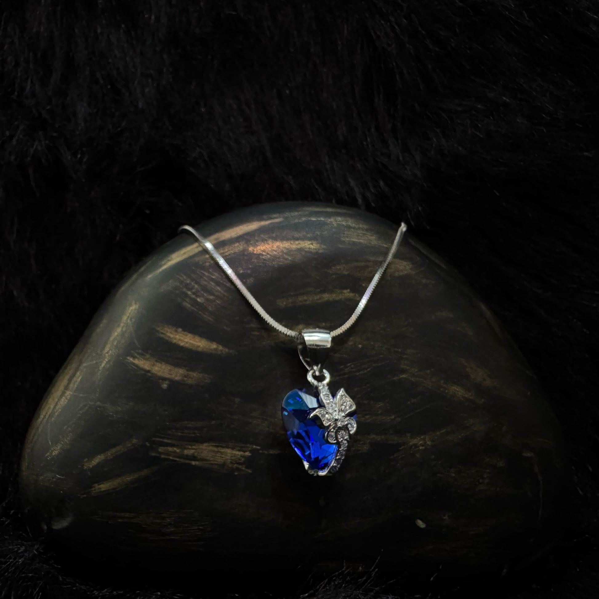 A silver necklace featuring a deep blue heart-shaped crystal pendant with a sparkling butterfly motif, displayed on a dark textured background.