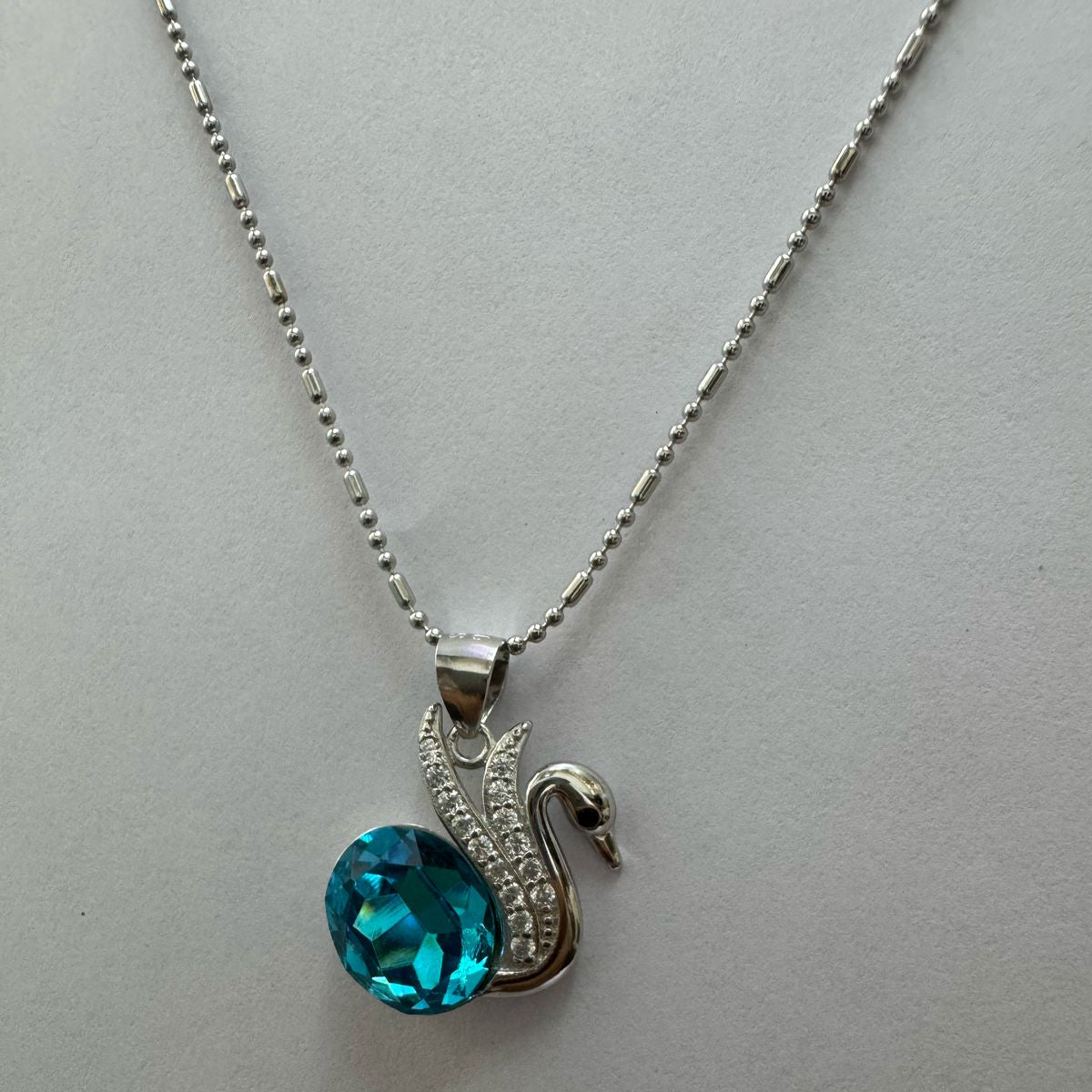 A silver necklace featuring an elegant swan motif adorned with cubic zirconia, holding a vivid blue oval-cut crystal, displayed against a textured black and gold background.