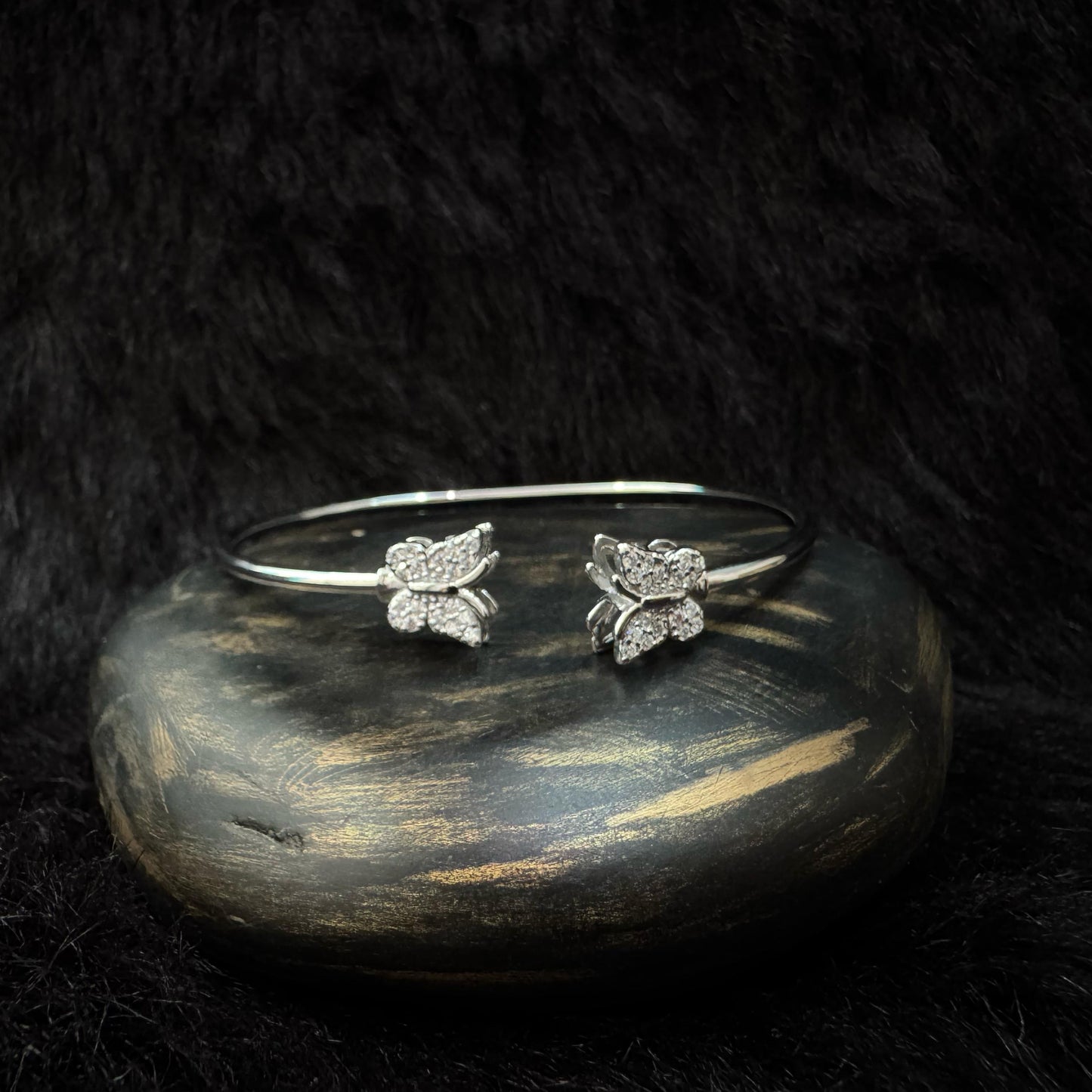 Elegant silver bangle bracelet with two butterfly-shaped ends adorned with sparkling cubic zirconia, displayed on a dark stone with a soft black background.