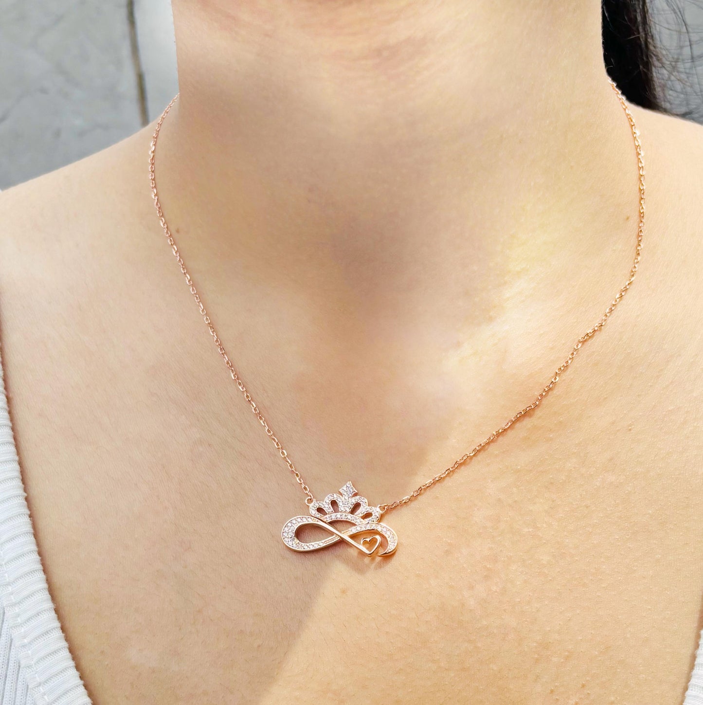 A rose-gold sterling silver infinity necklace featuring a crown, sparkling zirconia accents, and a heart design, worn elegantly on a woman's neck.