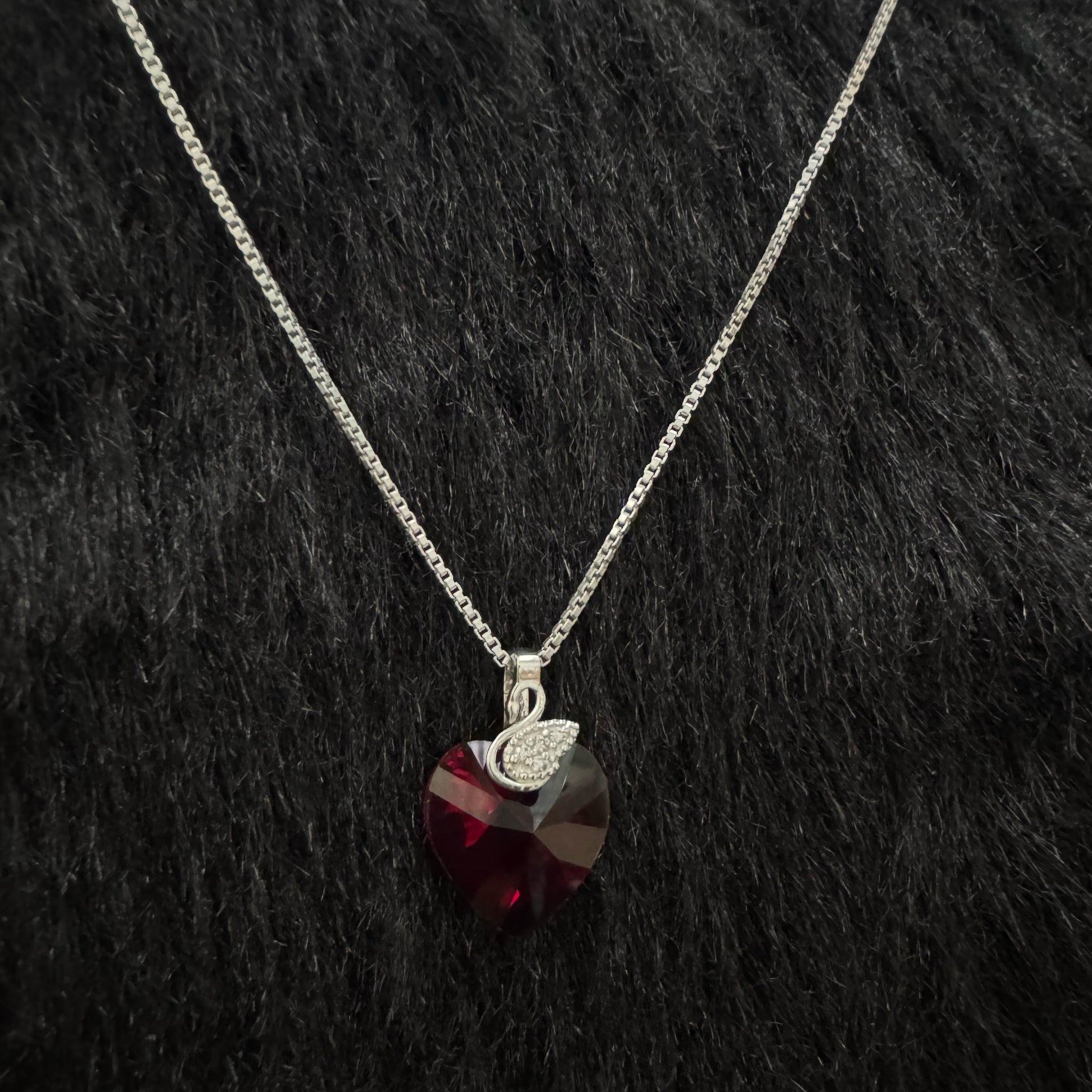 A sterling silver necklace featuring a deep red Swarovski heart-shaped crystal with a pavé cubic zirconia teardrop charm, elegantly displayed against a black textured background.
