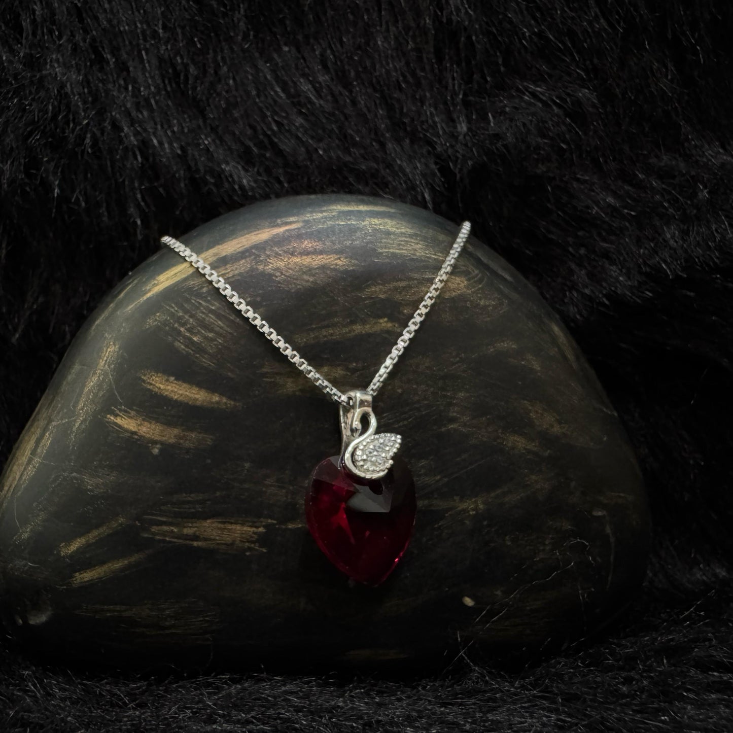 A sterling silver necklace featuring a deep red Swarovski heart-shaped crystal with a pavé cubic zirconia teardrop charm, elegantly displayed against a black textured background.