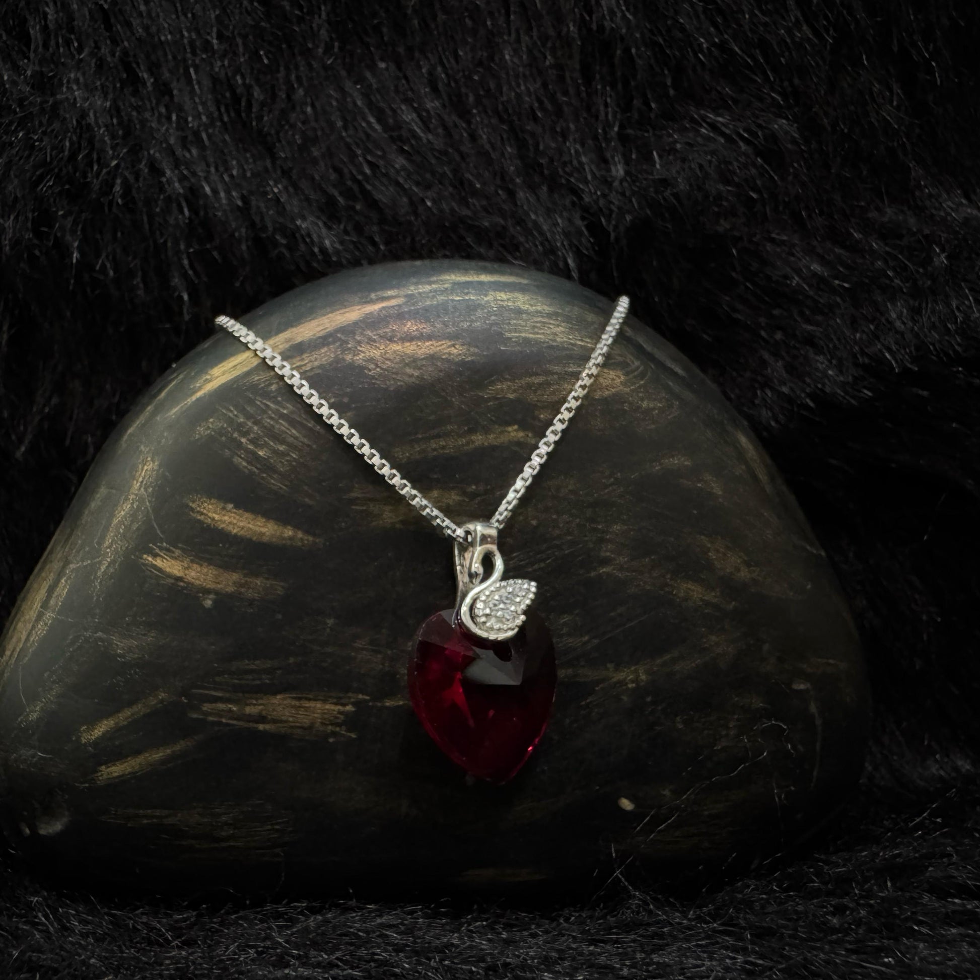 A sterling silver necklace featuring a deep red Swarovski heart-shaped crystal with a pavé cubic zirconia teardrop charm, elegantly displayed against a black textured background.