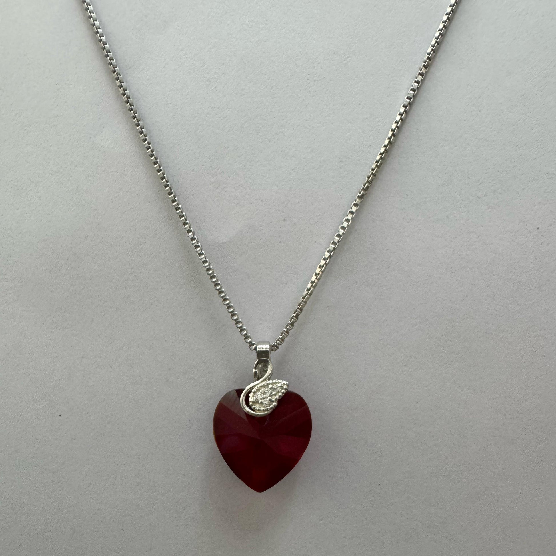 A sterling silver necklace featuring a deep red Swarovski heart-shaped crystal with a pavé cubic zirconia teardrop charm, elegantly displayed against a black textured background.