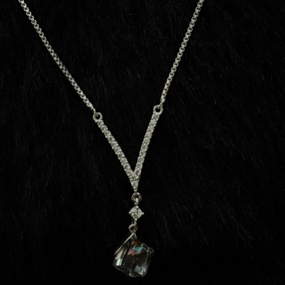 A silver rhinestone necklace featuring a bold cube-cut Swarovski crystal pendant, draped elegantly over a dark stone with a black background.