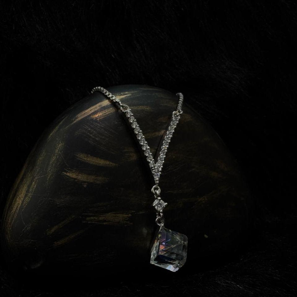 A silver rhinestone necklace featuring a bold cube-cut Swarovski crystal pendant, draped elegantly over a dark stone with a black background.