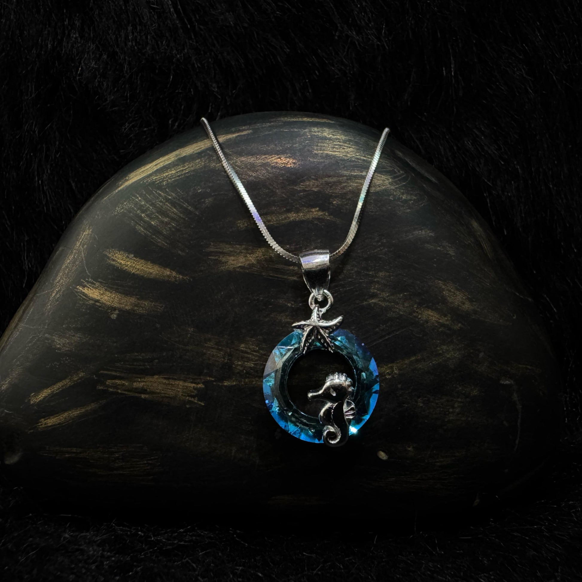 A silver necklace featuring a circular blue crystal pendant with a silver seahorse and starfish detail, displayed on a white background.