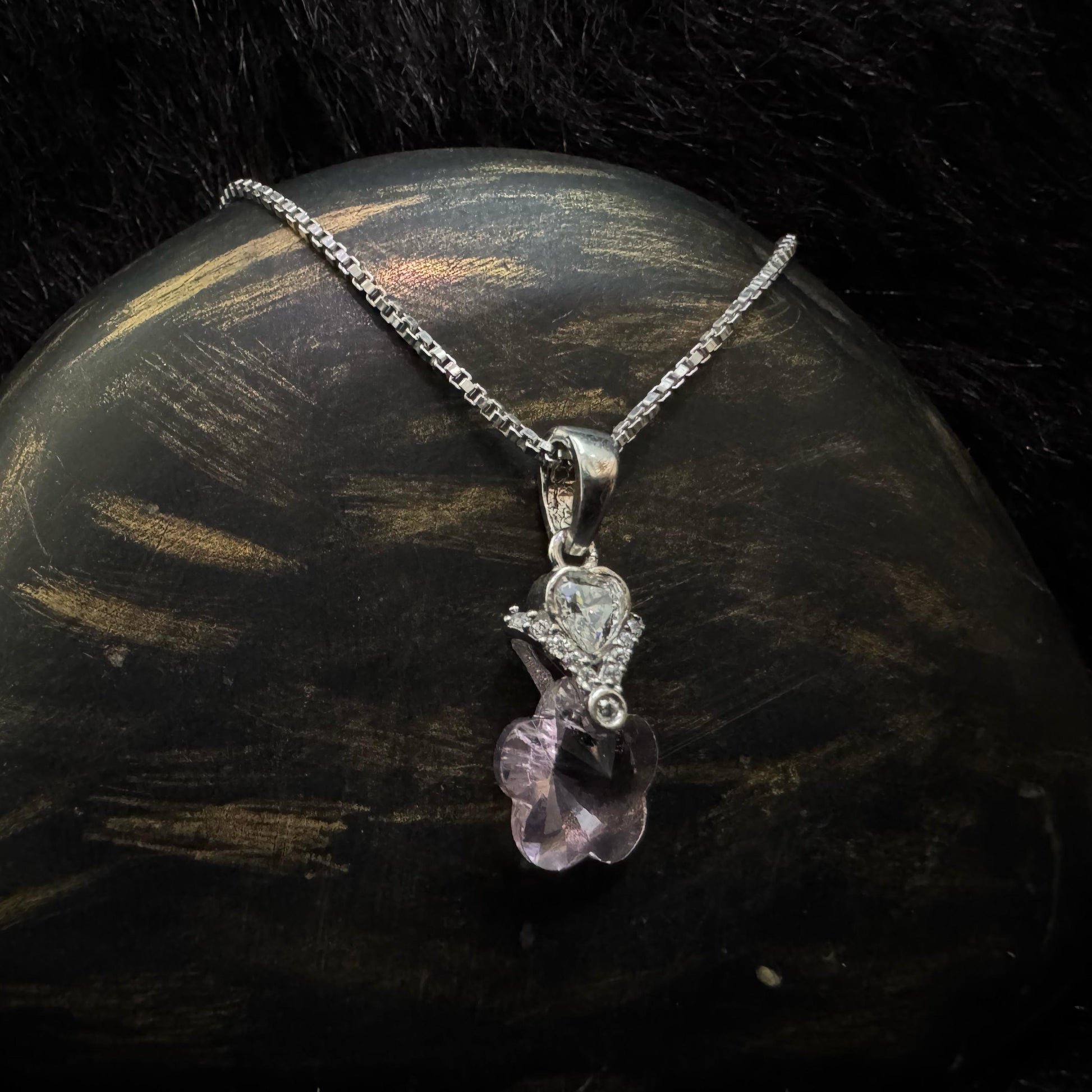 A sterling silver necklace with a pink heart-shaped crystal pendant, accented by a teardrop cubic zirconia and floral designs, displayed on a textured black and gold surface.