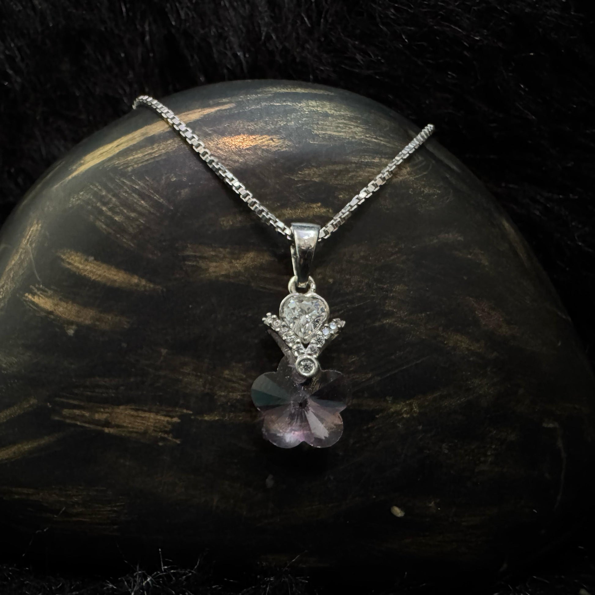 A sterling silver necklace with a pink heart-shaped crystal pendant, accented by a teardrop cubic zirconia and floral designs, displayed on a textured black and gold surface.