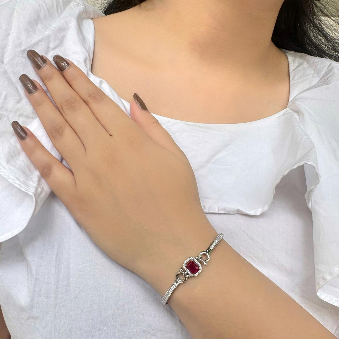 A hand wearing a silver bracelet with a central ruby-red stone and cubic zirconia halo, displayed on a white fabric background.