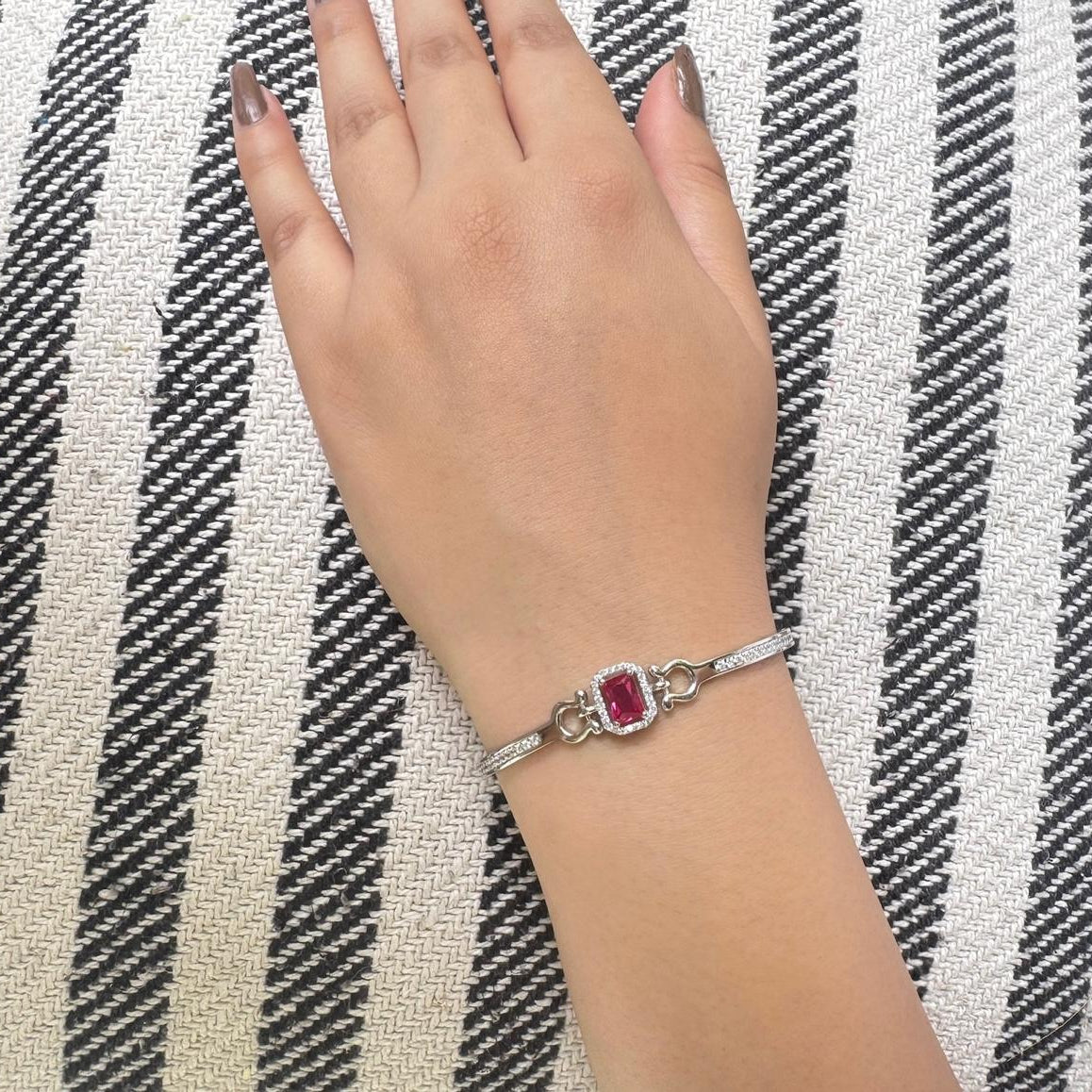 A hand wearing a silver bracelet with a central ruby-red stone and cubic zirconia halo, displayed on a white fabric background.