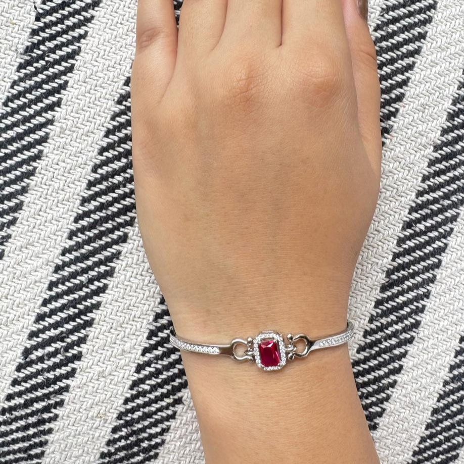 A hand wearing a silver bracelet with a central ruby-red stone and cubic zirconia halo, displayed on a white fabric background.