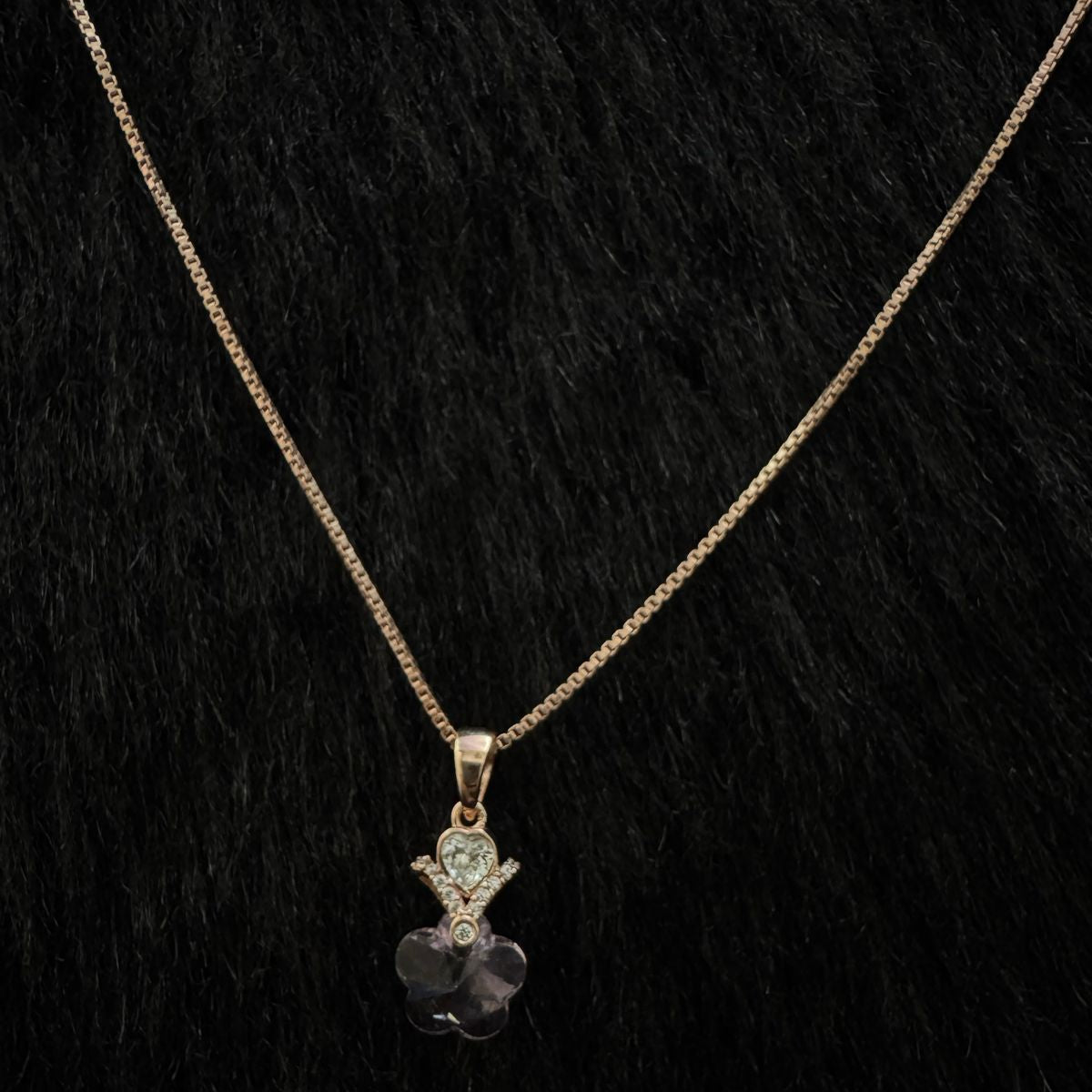 A rose gold necklace featuring a pendant of hands holding a heart above a clear faceted flower crystal, displayed elegantly on a dark stone with a black background.